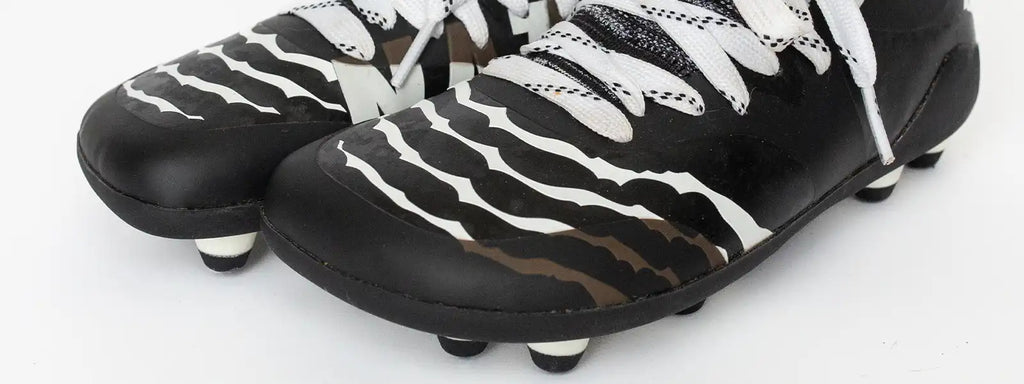 Close-up of Natur Athletics cleats designed for natural foot development and performance, featuring a sleek black and white design with stripes and cleat studs for optimal traction. These cleats are engineered to support growth, balance, and agility while respecting the foot's natural shape for young athletes.